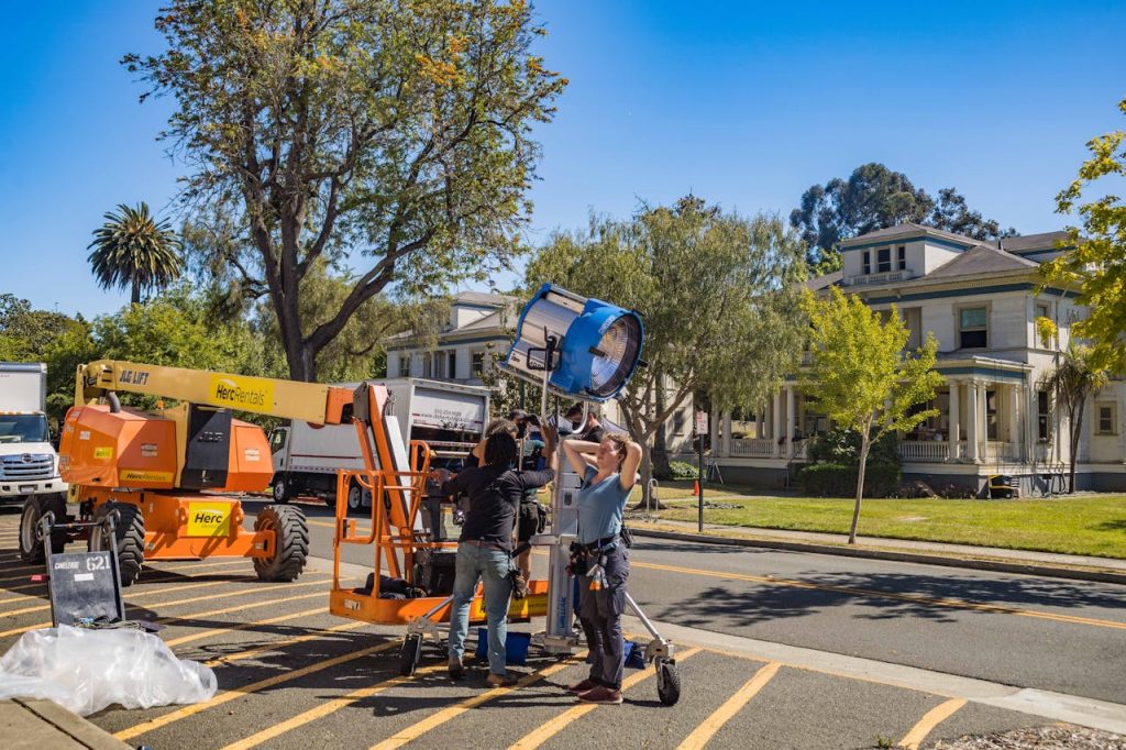 Film crew setting up lighting equipment outdoors with lift and tree backdrop.